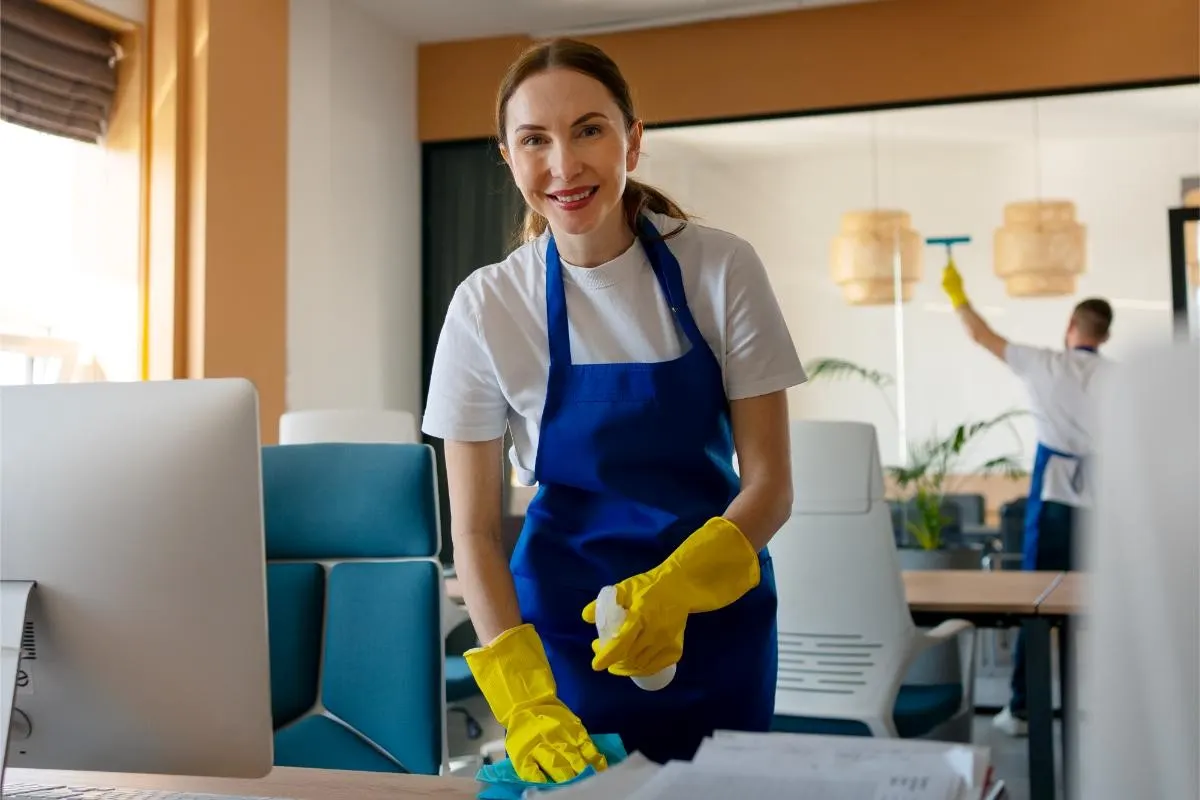 Smiling cleaner in blue apron and yellow gloves wiping an office desk while colleague cleans glass in background