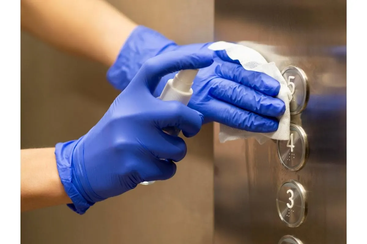 Gloved hands disinfecting elevator control panel buttons with sanitiser spray and wipe in a London building