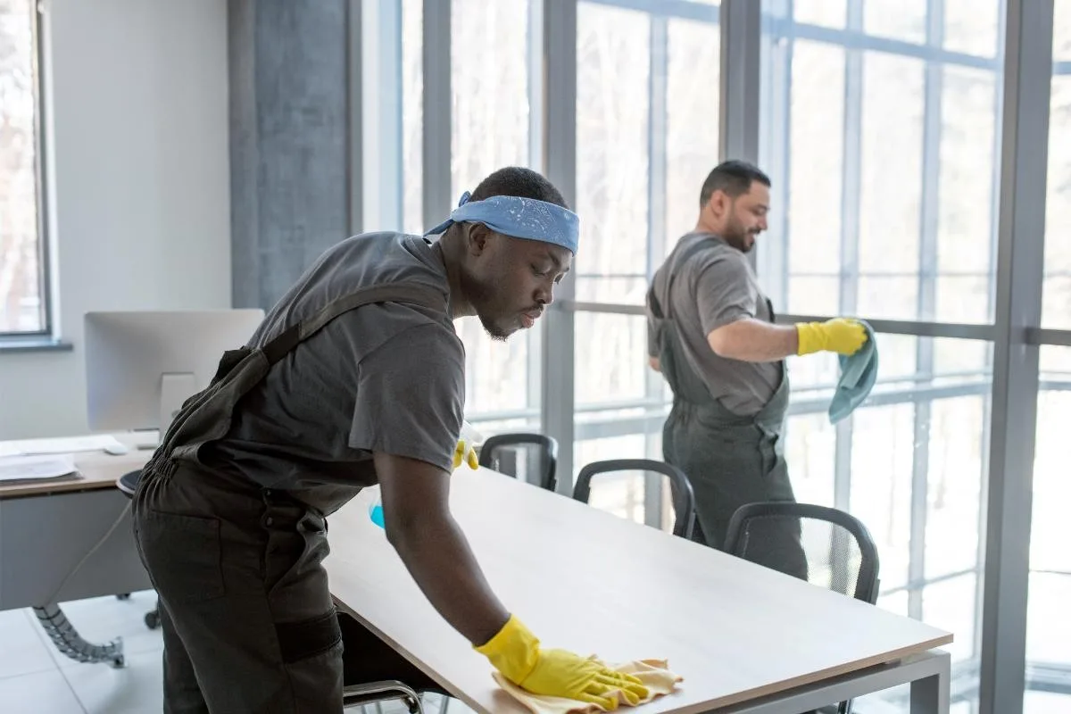 Two uniformed cleaners wiping tables and windows in a bright London office workspace